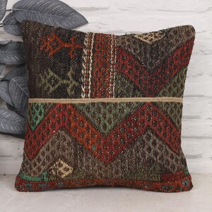 May include: Square decorative pillow with a geometric pattern in shades of brown, red, green, and beige. The pillow has a textured appearance and a horizontal beige stripe. The pillow is placed in front of a white brick wall.