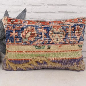 May include: Rectangular pillow with a vintage rug design. The pillow features a central section with a blue and red floral pattern, bordered by a beige, green, and purple striped section. The pillow is placed against a white brick wall.