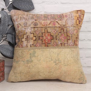 May include: A decorative pillow with a two-tone design. The top portion features a patterned, faded, multi-colored design with shades of brown, pink, and orange. The bottom half is a solid, light beige color. The pillow is placed against a white brick wall.