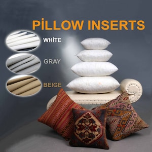 Puede incluir: Insertos de almohada en blanco, gris y beige. Los insertos están apilados uno encima del otro. Los insertos están en un sofá blanco con dos almohadas de tiro estampadas.