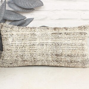 May include: Rectangular beige and brown patterned pillow. The pillow has a woven texture and is placed on a white surface. Decorative leaves and a small planter are in the background.