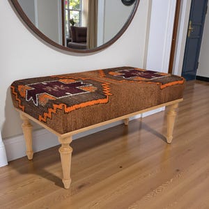 May include: A rectangular bench with a brown upholstered top featuring geometric patterns in orange and burgundy. The bench has light-coloured wooden legs. The bench is placed in front of a large round mirror.
