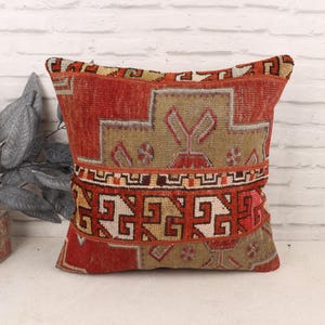 May include: A square, decorative pillow with a vintage, patterned design. The pillow features a red and beige color scheme with geometric and floral motifs. The pillow is likely made of woven fabric and adds a touch of rustic charm to any space.