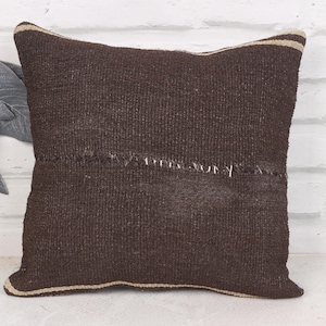 May include: A square, dark brown woven pillow with a beige border. The pillow has a stitched seam across the front. The pillow is made of a textured fabric and is placed against a white brick wall.