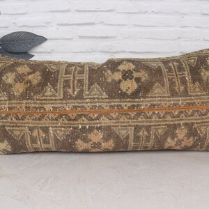 May include: A long, rectangular pillow with a brown and beige patterned fabric. The pillow has a woven design with geometric shapes and floral motifs. The fabric has a slightly distressed look, giving it a vintage feel.
