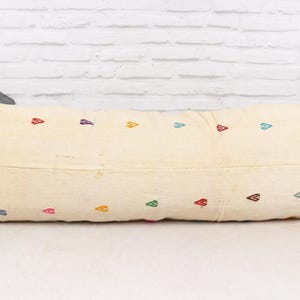 May include: Cream-colored lumbar pillow with colorful embroidered geometric shapes. The rectangular pillow has a stitched edge and is placed on a white surface. The pillow is a decorative item for a bed or sofa.