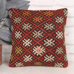 May include: Square decorative pillow with a geometric pattern. The pillow features a red background with a diamond pattern, and cream, orange, and yellow accents. The pillow is placed next to a decorative plant.