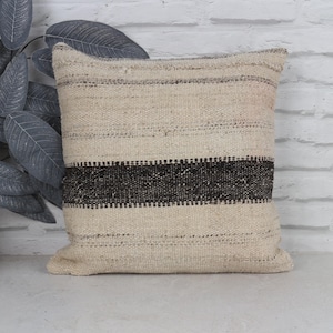 Peut inclure: Coussin carré beige et noir tissé. L'oreiller a une bande noire horizontale au centre et est fait d'un tissu texturé. Le coussin est posé sur une surface blanche.