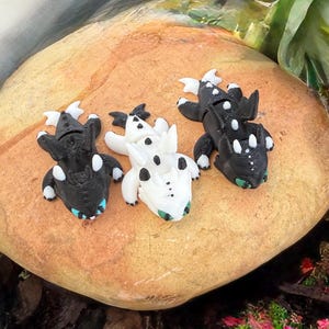 Puede incluir: Tres figuras de dragones en blanco y negro, con detalles en azul y verde, est&aacute;n dispuestas sobre una gran roca de color marr&oacute;n claro. Los dragones tienen detalles, incluyendo alas y manchas. La escena est&aacute; ambientada al aire libre con vegetaci&oacute;n de fondo.