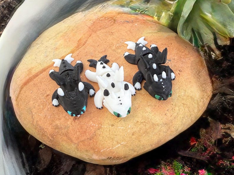 Puede incluir: Tres figuras de dragones en blanco y negro, con detalles en azul y verde, est&aacute;n dispuestas sobre una gran roca de color marr&oacute;n claro. Los dragones tienen detalles, incluyendo alas y manchas. La escena est&aacute; ambientada al aire libre con vegetaci&oacute;n de fondo.