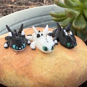 Puede incluir: Tres peque&ntilde;as figuras de dragones en blanco y negro, con ojos azules y verdes, descansan sobre una roca de color canela. Los dragones tienen detalles precisos y est&aacute;n colocados en una posici&oacute;n relajada. Una planta suculenta es visible en el fondo.