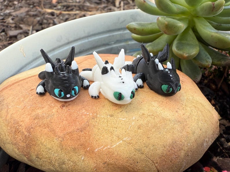 Puede incluir: Tres peque&ntilde;as figuras de dragones en blanco y negro, con ojos azules y verdes, descansan sobre una roca de color canela. Los dragones tienen detalles precisos y est&aacute;n colocados en una posici&oacute;n relajada. Una planta suculenta es visible en el fondo.