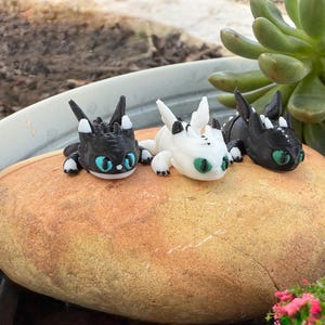 Puede incluir: Tres peque&ntilde;as figuras de dragones en negro, blanco y negro se exhiben sobre una gran roca de color marr&oacute;n claro. Los dragones tienen ojos grandes, redondos y turquesas y est&aacute;n en posici&oacute;n de descanso. El fondo incluye una planta suculenta verde y una maceta gris.