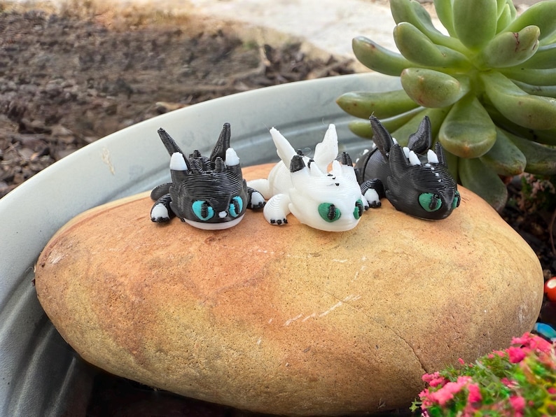 Puede incluir: Tres peque&ntilde;as figuras de dragones en negro, blanco y negro se exhiben sobre una gran roca de color marr&oacute;n claro. Los dragones tienen ojos grandes, redondos y turquesas y est&aacute;n en posici&oacute;n de descanso. El fondo incluye una planta suculenta verde y una maceta gris.