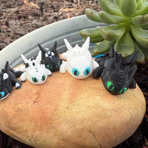 Puede incluir: Figuras de dragones impresas en 3D en varios colores, incluyendo negro, blanco y dise&ntilde;os moteados. Cada drag&oacute;n tiene ojos grandes con iris azules o verdes. Est&aacute;n colocados sobre una gran roca de color marr&oacute;n claro.