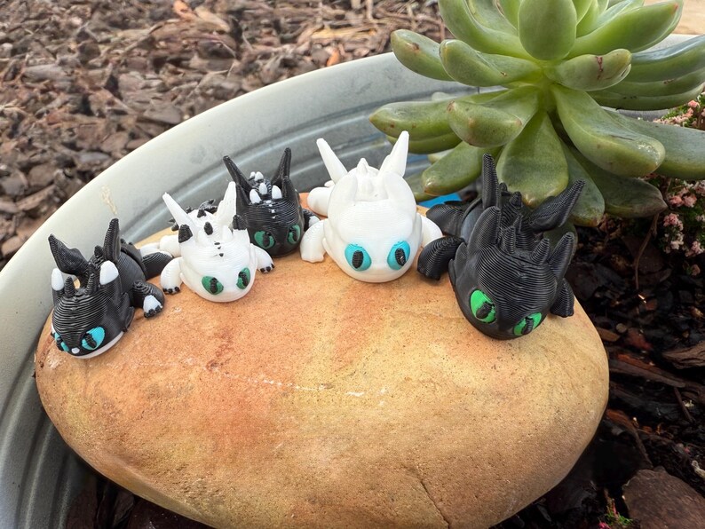 Puede incluir: Figuras de dragones impresas en 3D en varios colores, incluyendo negro, blanco y dise&ntilde;os moteados. Cada drag&oacute;n tiene ojos grandes con iris azules o verdes. Est&aacute;n colocados sobre una gran roca de color marr&oacute;n claro.
