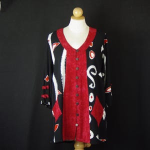 May include: A black tunic with a V-neckline and a central vertical red panel featuring a button-down closure. The garment displays a white and red abstract pattern throughout, with three-quarter length sleeves accented by small red fabric ties.
