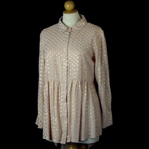 May include: A long-sleeved button-down shirt in a soft blush pink colour featuring a repeating pattern of metallic gold polka dots. The shirt has a classic collar, a front button placket, and a gathered waistline that creates a flared silhouette.