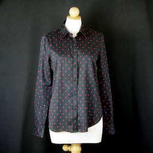 May include: A long-sleeved button-down shirt in black featuring a repeating pattern of small red polka dots. The shirt has a standard collar, a front button placket, and buttoned cuffs, displayed on a mannequin against a plain black background.