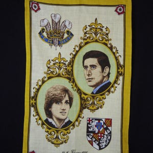 May include: A cream-coloured vintage handkerchief with a yellow border. It features portraits of Prince Charles and Princess Diana in ornate gold frames, a crest, and text. The date "26th February 1981" is visible, commemorating their engagement.