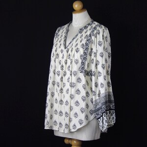 May include: A long-sleeved blouse in off-white featuring a repeating dark blue floral damask pattern. The garment has a V-neckline, gathered fabric at the shoulders, and elasticated cuffs on the sleeves, creating a loose and flowing silhouette.