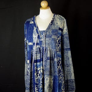 Peut inclure: Robe patchwork bleue et blanche à manches longues et col en V, présentée sur un mannequin. Le tissu présente divers motifs, notamment floraux et géométriques. La robe a un détail de cordon de serrage à l'encolure.