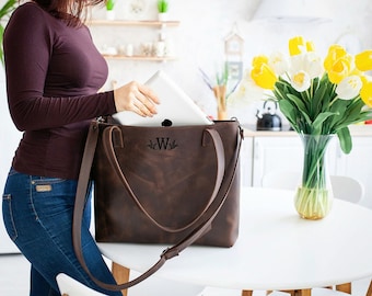 May include: A brown leather tote bag with a monogrammed "W" on the front. The bag has a long strap and is being used to carry a laptop.