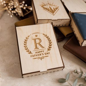 Puede incluir: Una caja rectangular de madera clara con tapa deslizante que presenta una corona de laurel grabada con l&aacute;ser, la letra R y el texto HAPPY FATHER'S DAY. Una cartera de cuero azul es visible dentro de una caja de madera abierta junto a otros objetos.