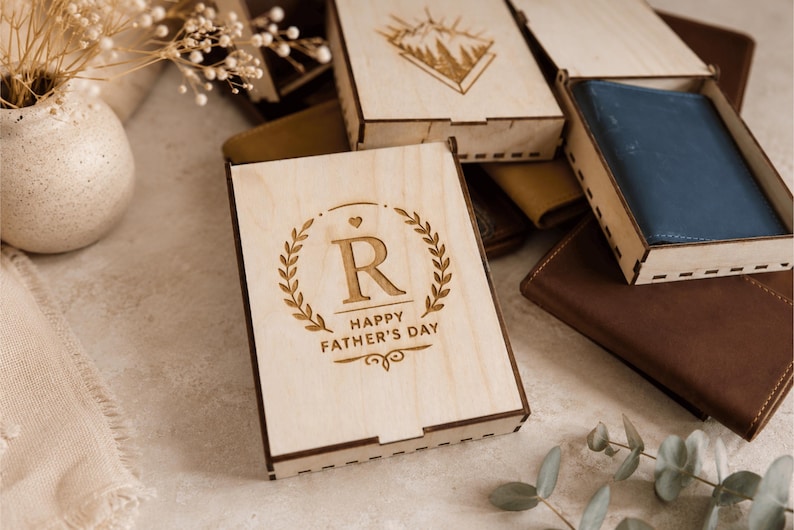 Puede incluir: Una caja rectangular de madera clara con tapa deslizante que presenta una corona de laurel grabada con l&aacute;ser, la letra R y el texto HAPPY FATHER'S DAY. Una cartera de cuero azul es visible dentro de una caja de madera abierta junto a otros objetos.