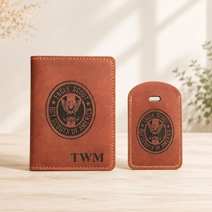 Puede incluir: Un portapasaportes de cuero marr&oacute;n y una etiqueta de equipaje a juego, ambos con un emblema negro grabado de Eagle Scout Boy Scouts of America. El portapasaportes est&aacute; personalizado con las letras negras TWM en la parte inferior central.