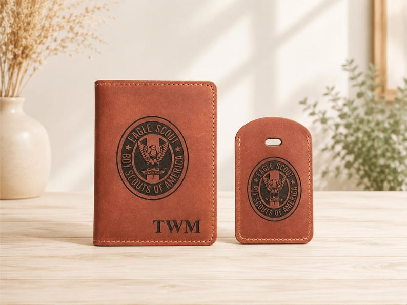 Puede incluir: Un portapasaportes de cuero marr&oacute;n y una etiqueta de equipaje a juego, ambos con un emblema negro grabado de Eagle Scout Boy Scouts of America. El portapasaportes est&aacute; personalizado con las letras negras TWM en la parte inferior central.