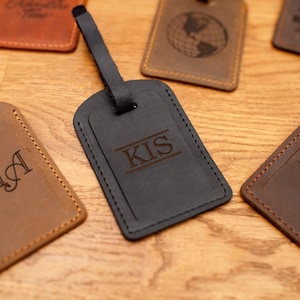 Puede incluir: Una colecci&oacute;n de etiquetas de equipaje rectangulares de cuero en varios tonos de marr&oacute;n y negro. Cada etiqueta cuenta con una correa de cuero y est&aacute; grabada con texto personalizado que incluye las letras KIA, KIS y KEA, junto con iconos de globos y viajes.