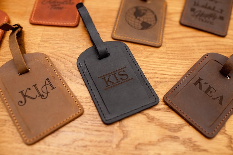 Puede incluir: Una colecci&oacute;n de etiquetas de equipaje rectangulares de cuero en varios tonos de marr&oacute;n y negro. Cada etiqueta cuenta con una correa de cuero y est&aacute; grabada con texto personalizado que incluye las letras KIA, KIS y KEA, junto con iconos de globos y viajes.