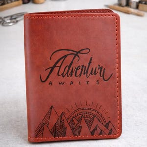 Puede incluir: Un portapasaportes rectangular de color &oacute;xido con un grabado marr&oacute;n oscuro. El texto "Adventure AWAITS" est&aacute; centrado sobre una ilustraci&oacute;n gr&aacute;fica de picos monta&ntilde;osos y un sol naciente con l&iacute;neas radiantes.