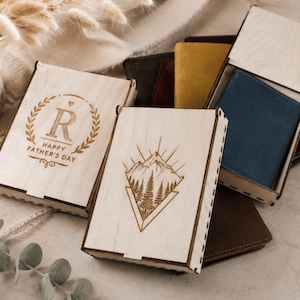 Puede incluir: Varias cajas rectangulares de madera clara con tapas deslizantes, algunas con dise&ntilde;os grabados a l&aacute;ser que incluyen un paisaje de monta&ntilde;a y una corona de laurel con la letra R y el texto HAPPY FATHER'S DAY, que contienen carteras de cuero.