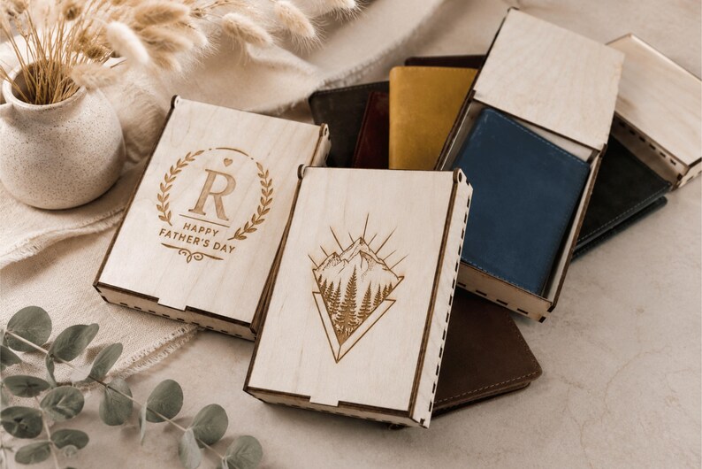 Puede incluir: Varias cajas rectangulares de madera clara con tapas deslizantes, algunas con dise&ntilde;os grabados a l&aacute;ser que incluyen un paisaje de monta&ntilde;a y una corona de laurel con la letra R y el texto HAPPY FATHER'S DAY, que contienen carteras de cuero.
