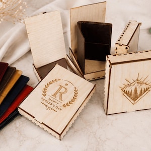 Puede incluir: Una colecci&oacute;n de cajas rectangulares de madera con dise&ntilde;os grabados a l&aacute;ser, incluyendo una con una escena de monta&ntilde;a y otra con la letra R y el texto HAPPY FATHER'S DAY. Varias carteras de cuero plegadas de varios colores est&aacute;n apiladas.