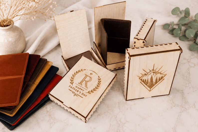 Puede incluir: Una colecci&oacute;n de cajas rectangulares de madera con dise&ntilde;os grabados a l&aacute;ser, incluyendo una con una escena de monta&ntilde;a y otra con la letra R y el texto HAPPY FATHER'S DAY. Varias carteras de cuero plegadas de varios colores est&aacute;n apiladas.
