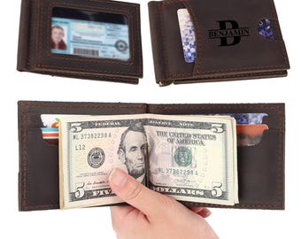 May include: A dark brown leather money clip wallet featuring a clear ID window, multiple card slots, and a metal money clip. The exterior is personalized with the name Benjamin and a large letter B. It holds several cards and a five-dollar bill.