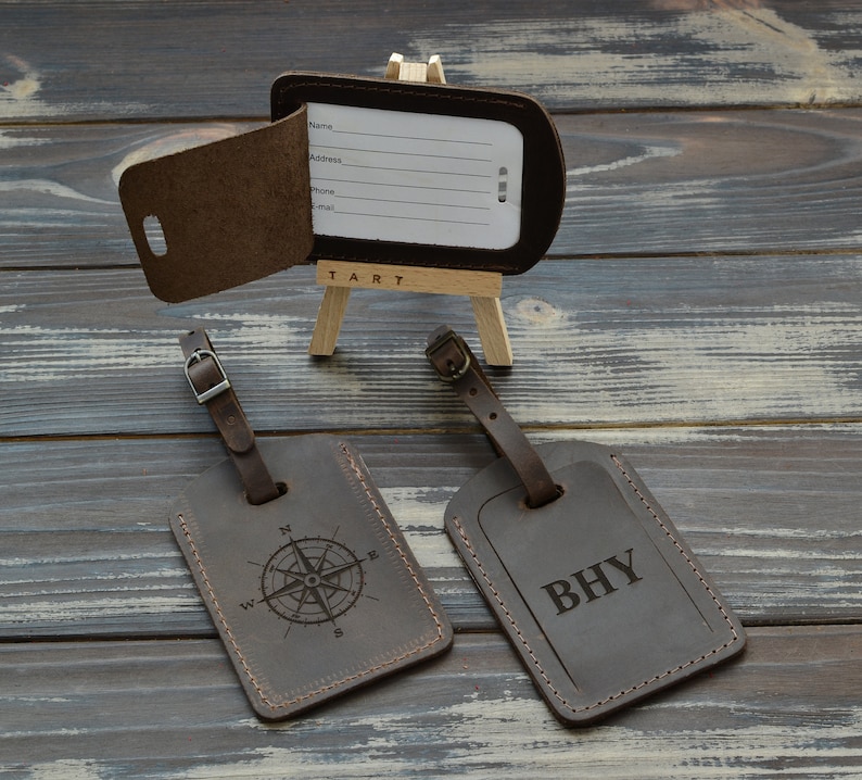 Puede incluir: Tres etiquetas de equipaje de cuero marr&oacute;n oscuro con correas ajustables y hebillas de metal. Una etiqueta est&aacute; abierta, revelando una tarjeta blanca con l&iacute;neas para Name, Address, Phone y E-mail. Las otras dos tienen grabados: una rosa de los vientos y las letras BHY.