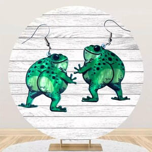 May include: A pair of frog-shaped earrings. Each earring features a green frog with black spots, a smiling face, and outstretched arms. The earrings are suspended from silver-colored fish hook ear wires. The frogs are set against a white wooden background.