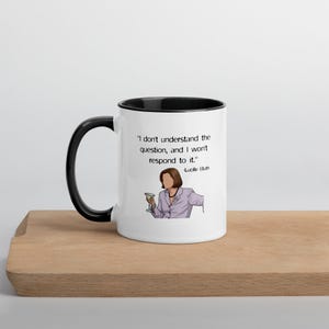 Può includere: Una tazza in ceramica bianca con manico e bordo interno neri. La tazza presenta un'illustrazione grafica di una persona con un blazer viola chiaro che tiene in mano un bicchiere da martini, accompagnata dal testo: I don't understand the question, and I won't respond to it. -Lucille Bluth.
