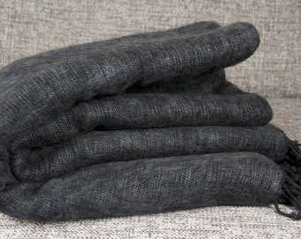 Pode incluir: Um cachecol dobrado, cinza escuro, com uma aparência macia e texturizada. O cachecol é feito de um material tecido e tem um detalhe de franjas na extremidade. O cachecol é dobrado sobre si mesmo, criando um efeito em camadas.