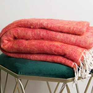 Op de afbeelding: Een gevouwen koraalkleurige deken met een geweven textuur en crèmekleurige franjes ligt op een groene fluwelen kruk met zilveren poten. De deken ziet er zacht en uitnodigend uit.