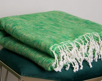 Pode incluir: Uma manta verde dobrada com franjas brancas é exibida sobre um banco estofado verde escuro com pernas prateadas. A manta tem uma aparência texturizada e é um tom vibrante de verde.