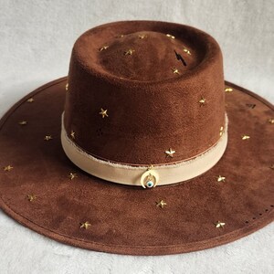 May include: A brown suede hat with a wide brim and a cream colored band. The hat is decorated with gold stars and a gold crescent moon charm with a blue evil eye.
