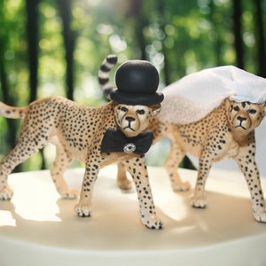 May include: A wedding cake topper featuring two cheetah figurines. The groom cheetah wears a black top hat and bow tie, while the bride cheetah wears a white veil.