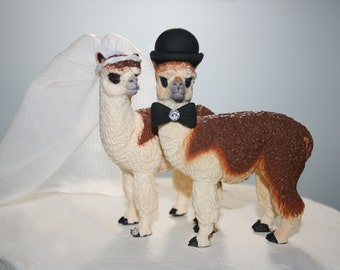 May include: A white alpaca wearing a white veil and a brown alpaca wearing a black top hat and a black bow tie with a rhinestone. The alpaca couple is standing on a white cake.