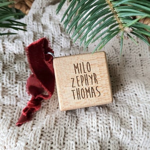May include: A wooden cube ornament with the names "Milo Zephyr Thomas" engraved on it. The ornament is hanging from a red ribbon and is resting on a white knitted blanket.
