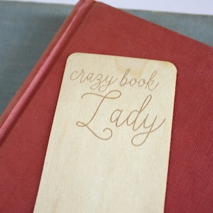 May include: A wooden bookmark with the text "crazy book Lady" engraved on it. The bookmark is light brown and has a natural wood grain.